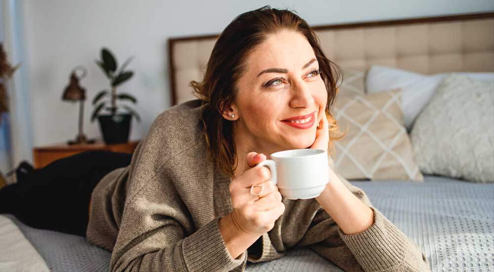 Mulher madura deitada em uma cama, segurando uma xícara e sorrindo, pois se sente bem com a reposição hormonal na menopausa da Lybe Clinic